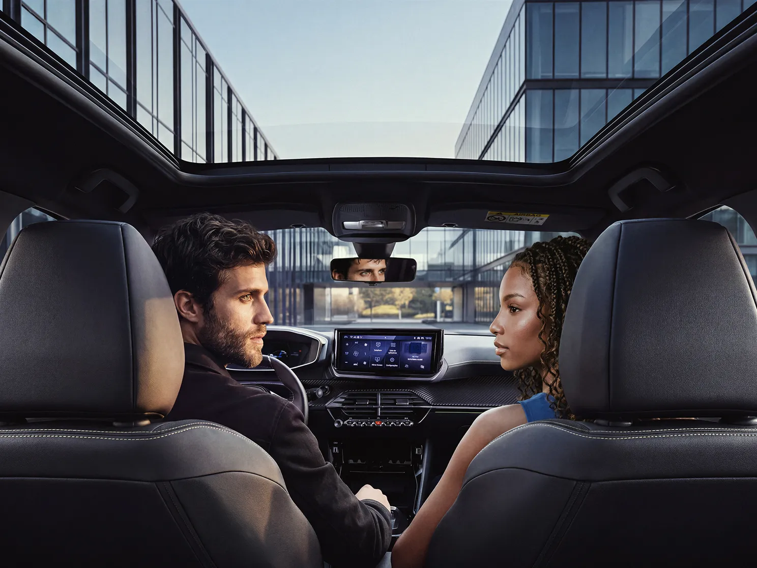 Homem e mulher conversam dentro de um Peugeot, vistos dos bancos traseiros, com painel multimídia ao centro. Casal no interior de um Peugeot.