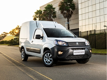 Peugeot Partner Rapid branco circula por via urbana, com edifício envidraçado e árvores ao fundo. Peugeot Partner Rapid em via urbana.