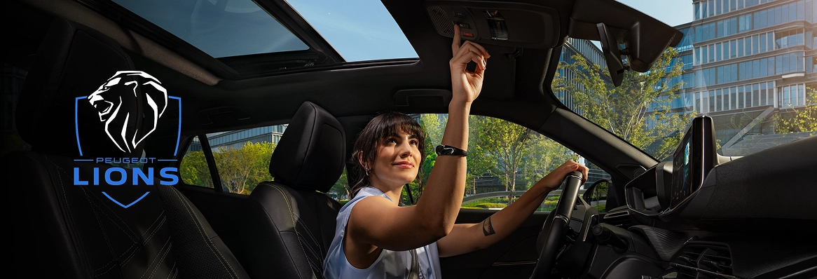 Interior de um Peugeot com mulher no banco do motorista com uma mão no volante e outra acionando botão no teto, com teto panorâmico aberto, painel e central multimídia visíveis e logo Peugeot Lions sobreposto. Peugeot Lions - Programa de relacionamento